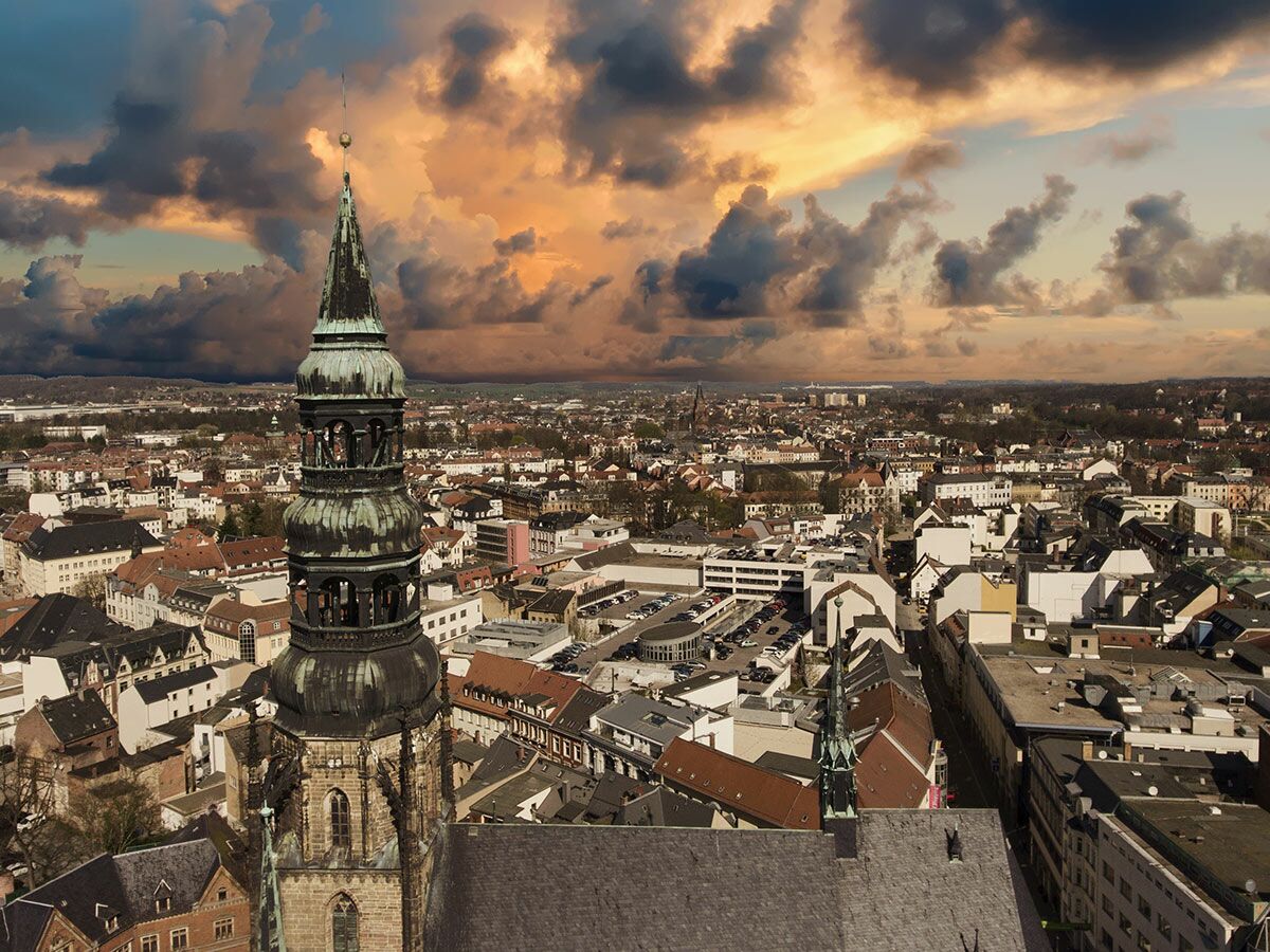 Luftbild einer europäischen Stadt mit markantem historischen Kirchturm unter einem spektakulären Sonnenuntergangshimmel.