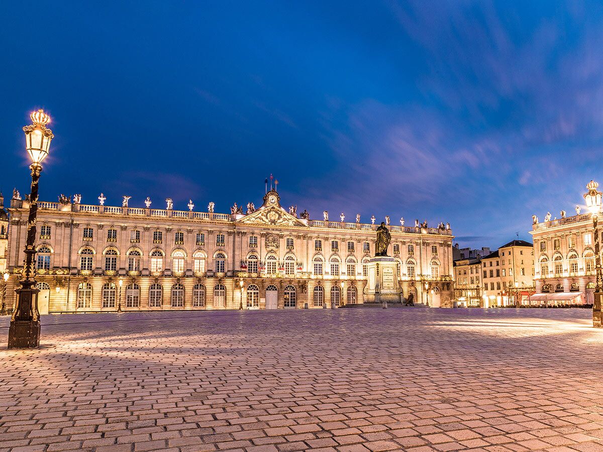 Place Stanislas in Nancy, Frankreich, mit dem abends beleuchteten Hôtel de Ville, umgeben von eleganten Barockgebäuden unter blauem Himmel.