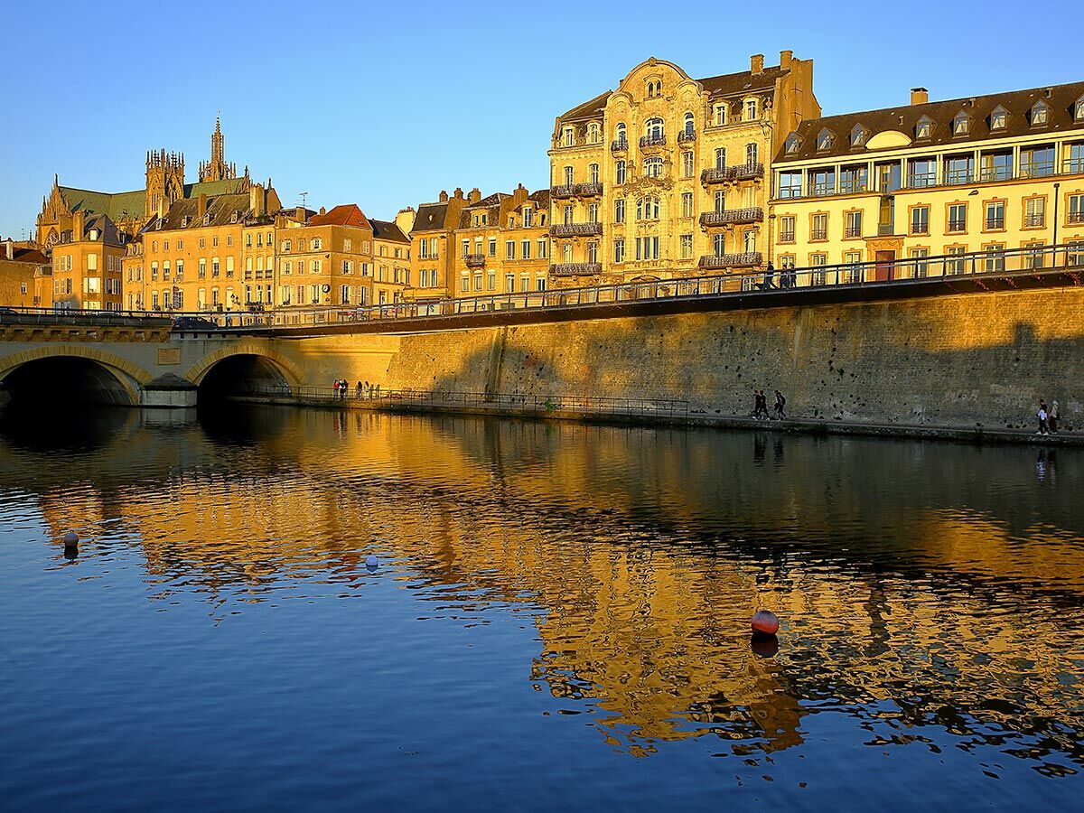 Metz: Historische Gebäude und Kathedrale spiegeln sich im Mosel-Fluss bei Sonnenuntergang; Menschen spazieren am Ufer.
