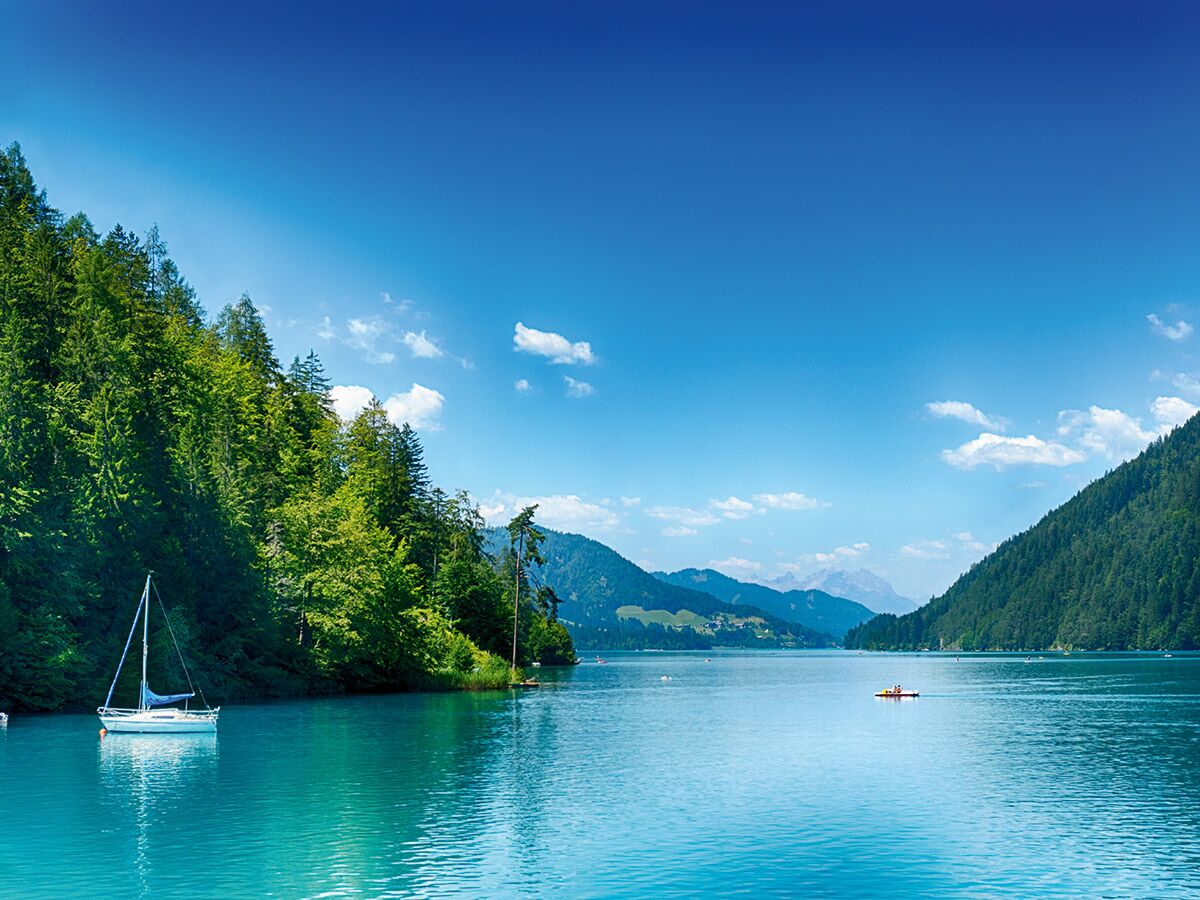 Idyllischer türkiser Bergsee mit Segelboot, umgeben von bewaldeten Bergen unter blauem Himmel und weißen Wolken.