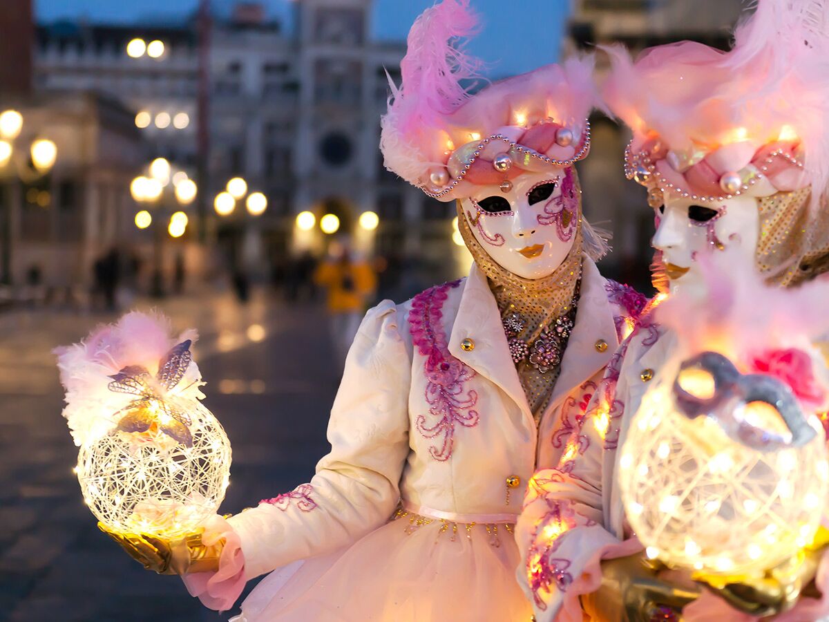Zwei Personen in prächtigen, pink-weißen venezianischen Karnevalsmasken und leuchtenden Kostümen halten glühende Laternen am Abend.