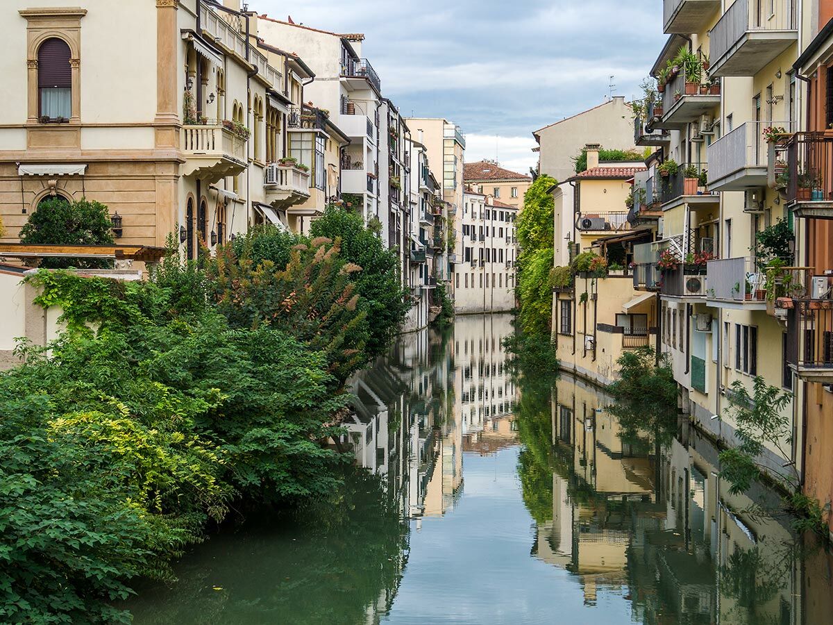 Blick auf einen malerischen Kanal in einer italienischen Stadt, gesäumt von historischen Gebäuden und üppigem Grün, die sich im Wasser spiegeln.