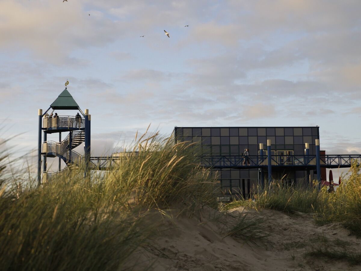 Moderner Aussichtsturm mit Wendeltreppe und verbundenem Gebäude, umgeben von Sanddünen mit Strandhafer unter bewölktem Himmel.