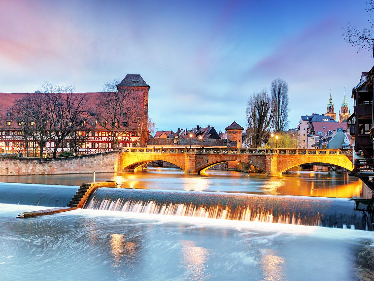 Blick auf Nürnbergs Altstadt am Abend: Weinstadel, Henkersteg-Brücke und Pegnitz mit Wehr bei Dämmerung, beleuchtet.