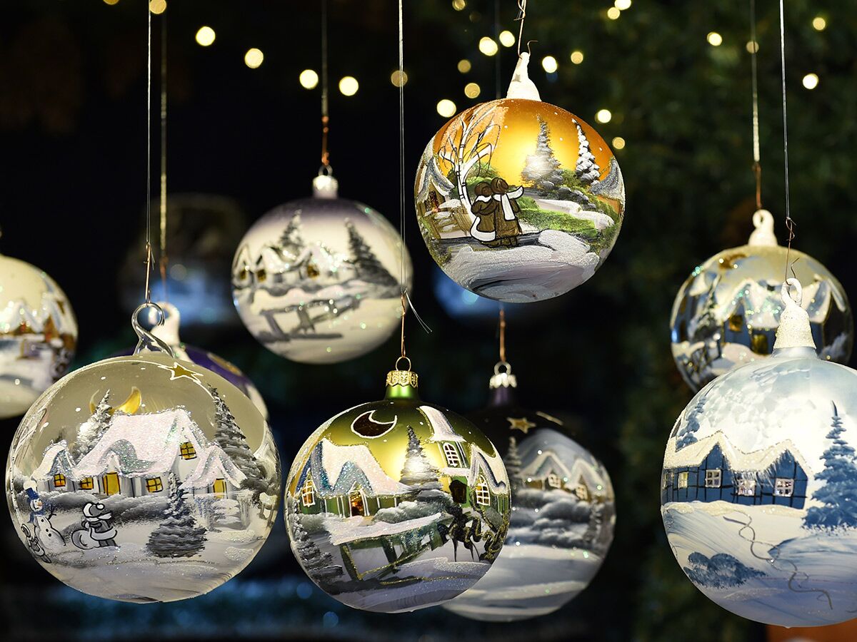 Nahaufnahme von handbemalten Weihnachtskugeln, die schneebedeckte Winterdörfer und Bäume zeigen, warme Lichter im unscharfen Hintergrund.