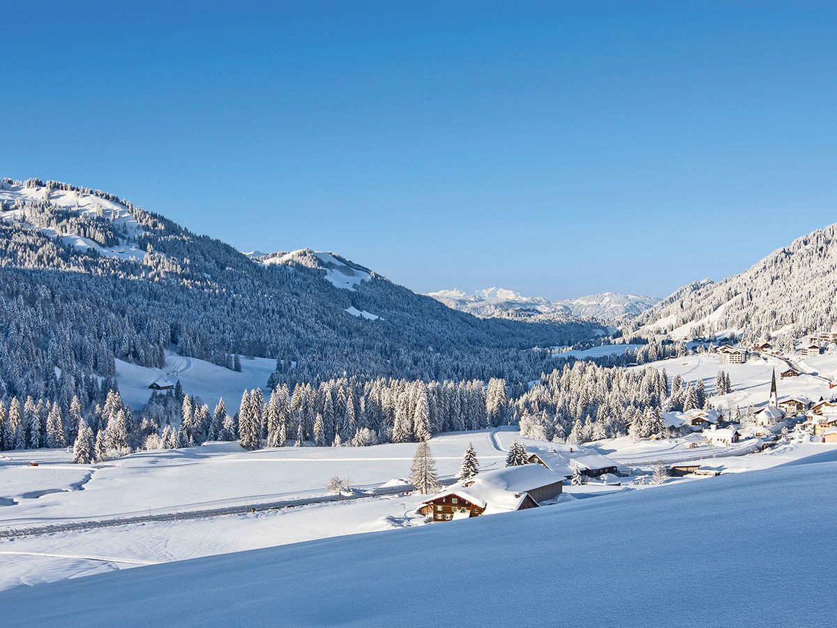 Idyllische Alpen-Winterlandschaft mit verschneiten Bergen, Tannenwäldern, sonnigem Tal, Dorf und Kirche unter blauem Himmel.