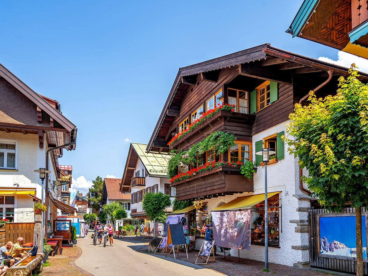 Eine idyllische Straße in Garmisch-Partenkirchen mit traditionellen Häusern, Blumenkästen und Menschen an einem sonnigen Sommertag.
