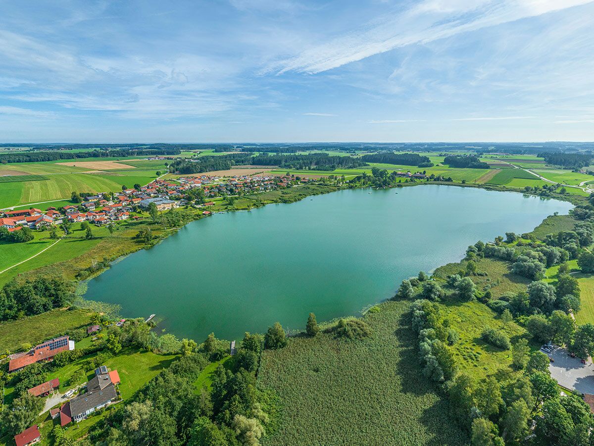 Luftaufnahme eines grün-blauen Sees mit angrenzendem Dorf, Feldern, Wäldern unter blauem Himmel.