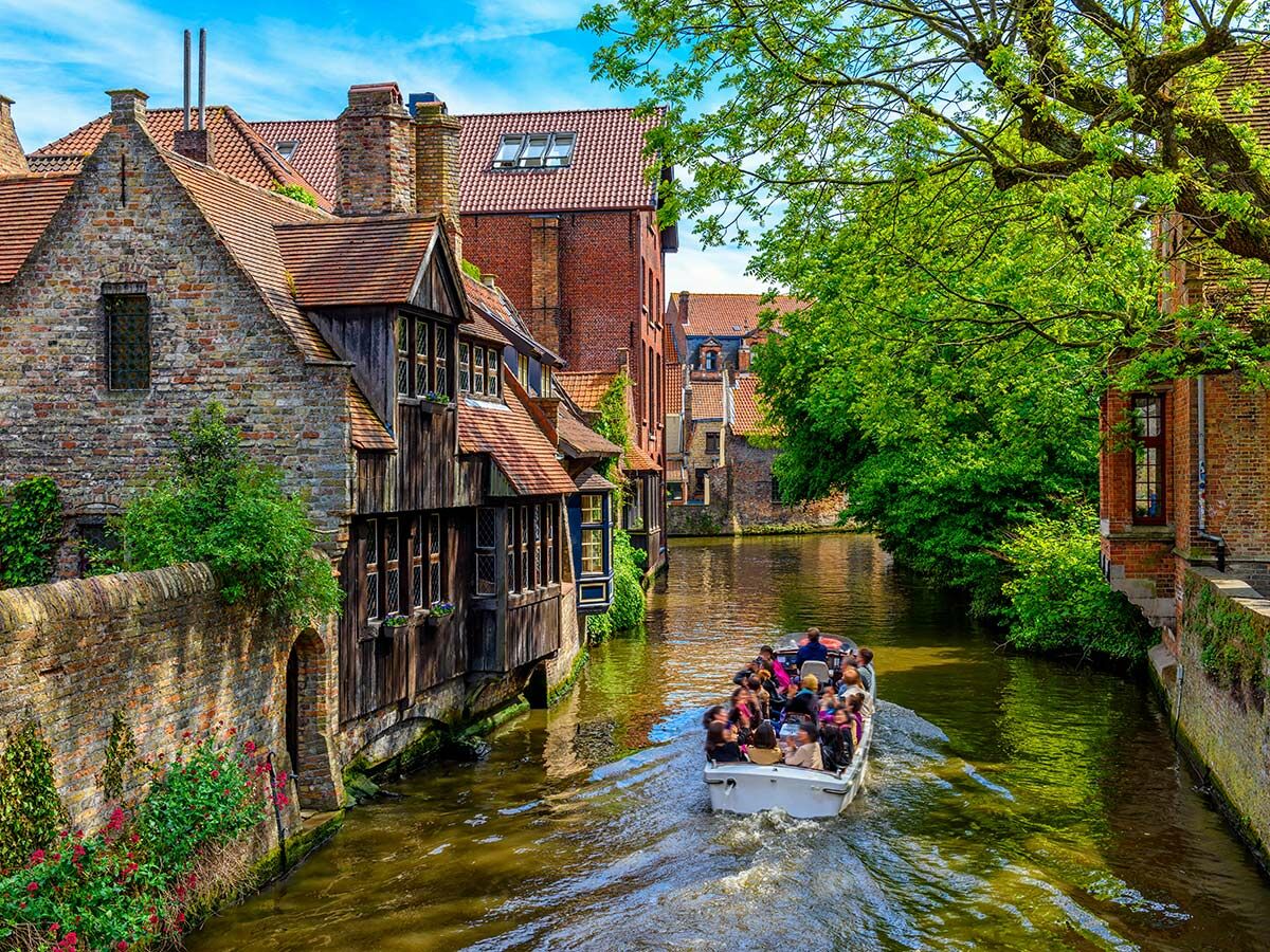 Touristenboot auf einem Kanal in Brügge, vorbei an alten Backsteinhäusern und grünen Bäumen unter blauem Himmel.