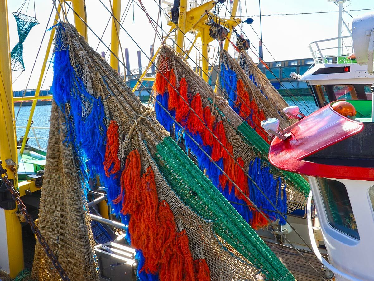 Farbenfrohe Fischernetze in Blau, Orange, Grün und Beige hängen an einem Fischerboot im Hafen, mit gelben Masten und dem Meer.