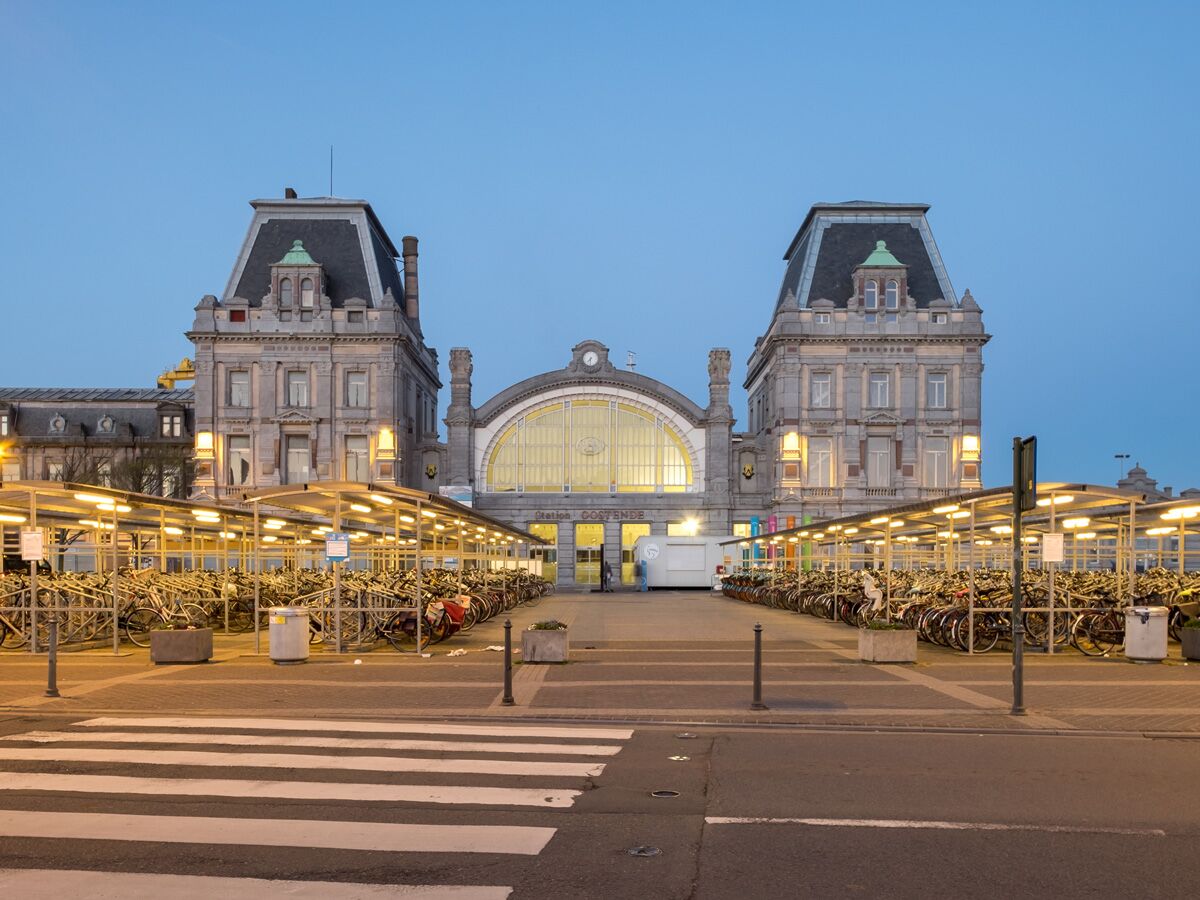Historischer Bahnhof Oostende bei Dämmerung mit vielen Fahrrädern unter beleuchteten Unterständen und Zebrastreifen.