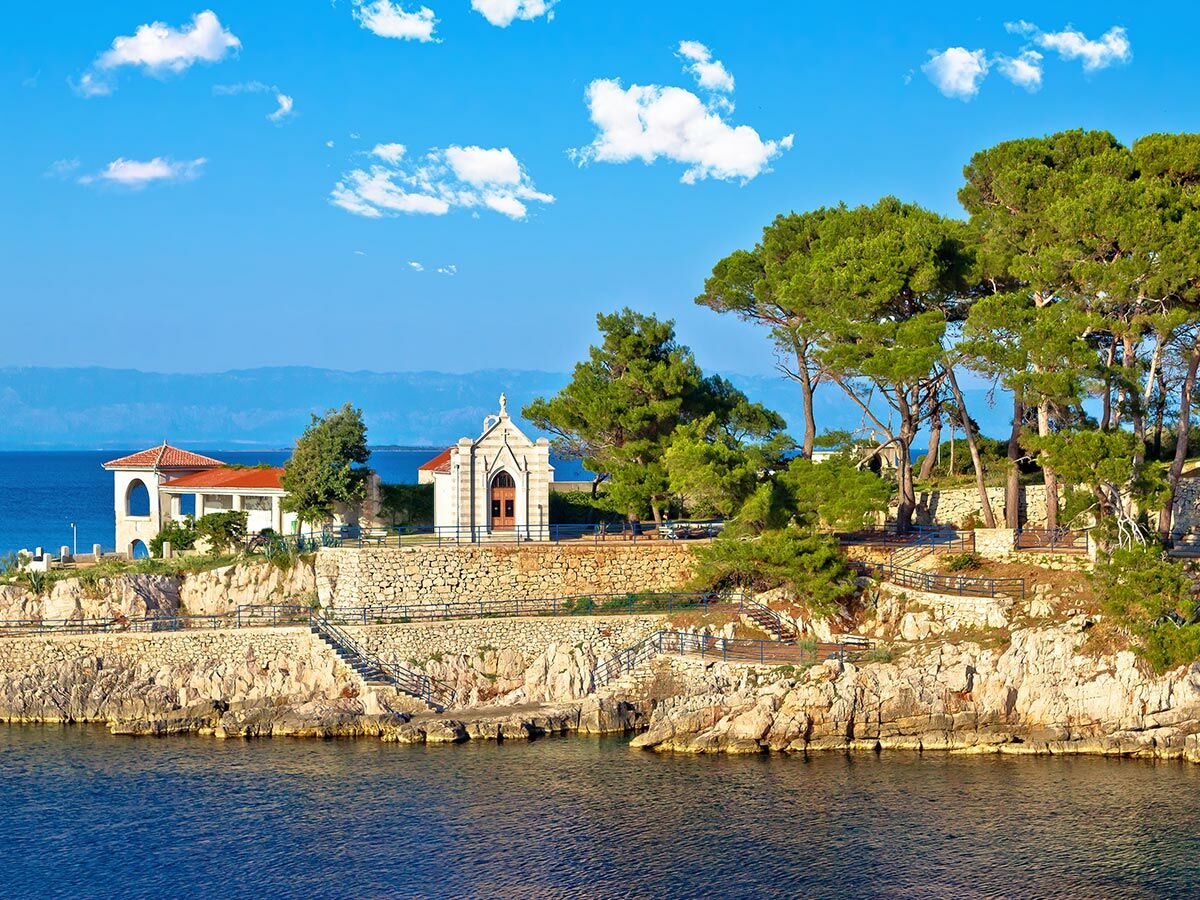 Panoramablick auf die sonnige Adriaküste mit weißer Kapelle, einem Haus, grünen Pinien und Felsen am blauen Meer unter blauem Himmel.