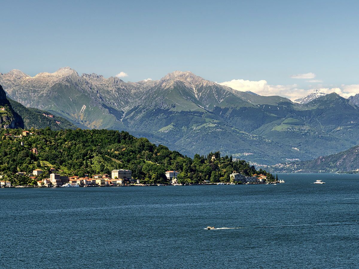 Atemberaubende Landschaft des Comer Sees mit charmantem Dorf am Ufer und majestätischen Bergen im Hintergrund unter blauem Himmel.