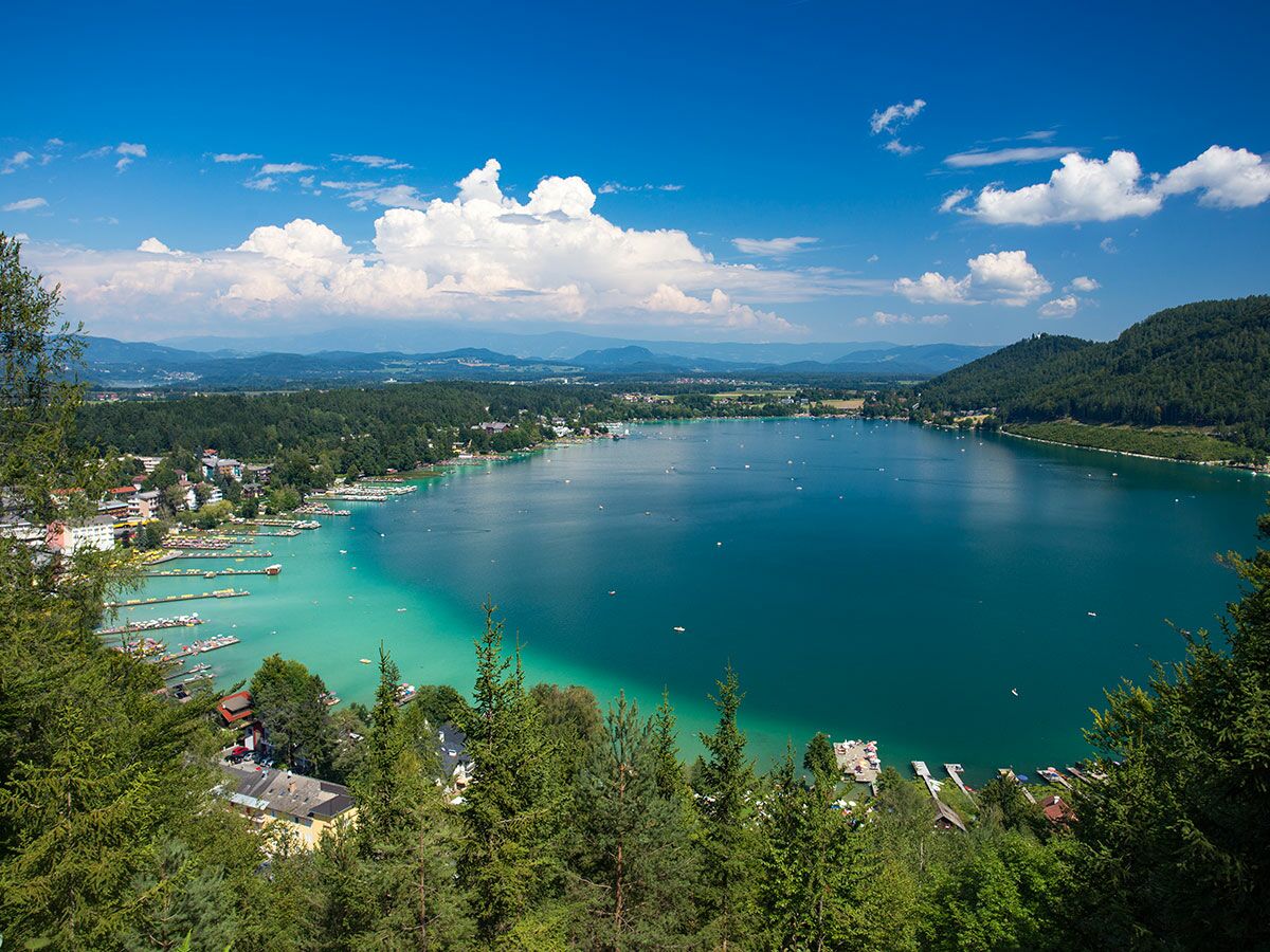 Panoramablick auf den türkisen Wörthersee in Kärnten, umgeben von grünen Wäldern, Häusern und Bergen unter blauem Himmel.