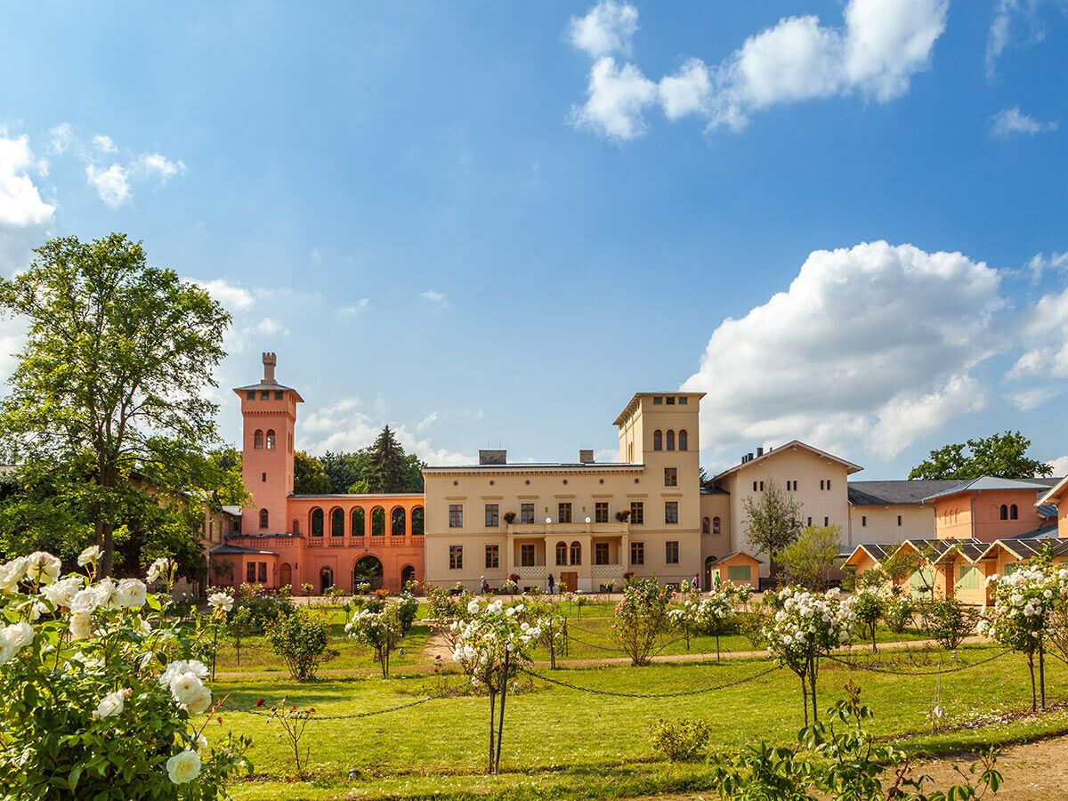 Malerisches Schlossgebäude mit rosafarbenem Turm und Bögen in einem blühenden Rosengarten unter blauem Himmel an einem sonnigen Tag.