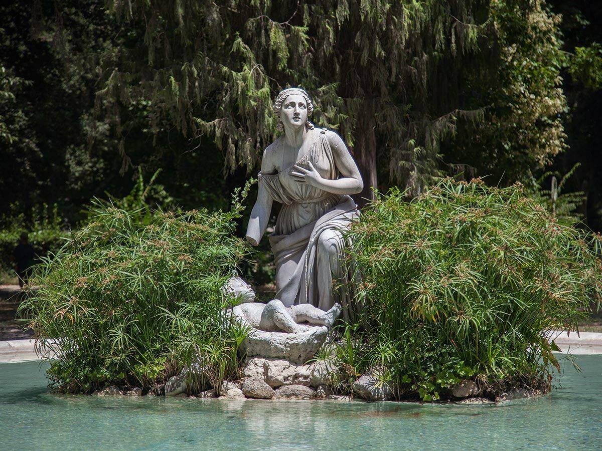 Klassische Marmorstatue einer Frau und eines Kindes in einem Teich, umgeben von grünen Pflanzen, in einem sonnigen Park.