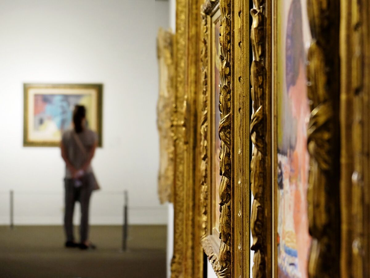 Eine verschwommene Person betrachtet ein Gemälde in einer Kunstgalerie, mit detailreichen goldenen Bilderrahmen im Vordergrund.