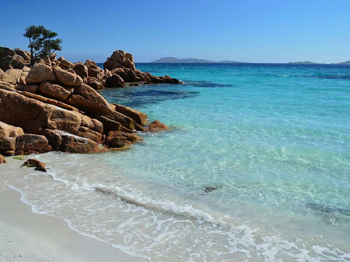 Idyllischer Strand in Sardinien mit großen roten Felsen, weißem Sand und kristallklarem, türkisfarbenem Wasser unter blauem Himmel.