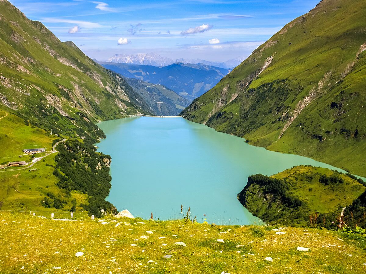 Ein türkiser Stausee eingebettet in eine grüne Berglandschaft mit Tälern und einem Damm unter blauem Himmel.