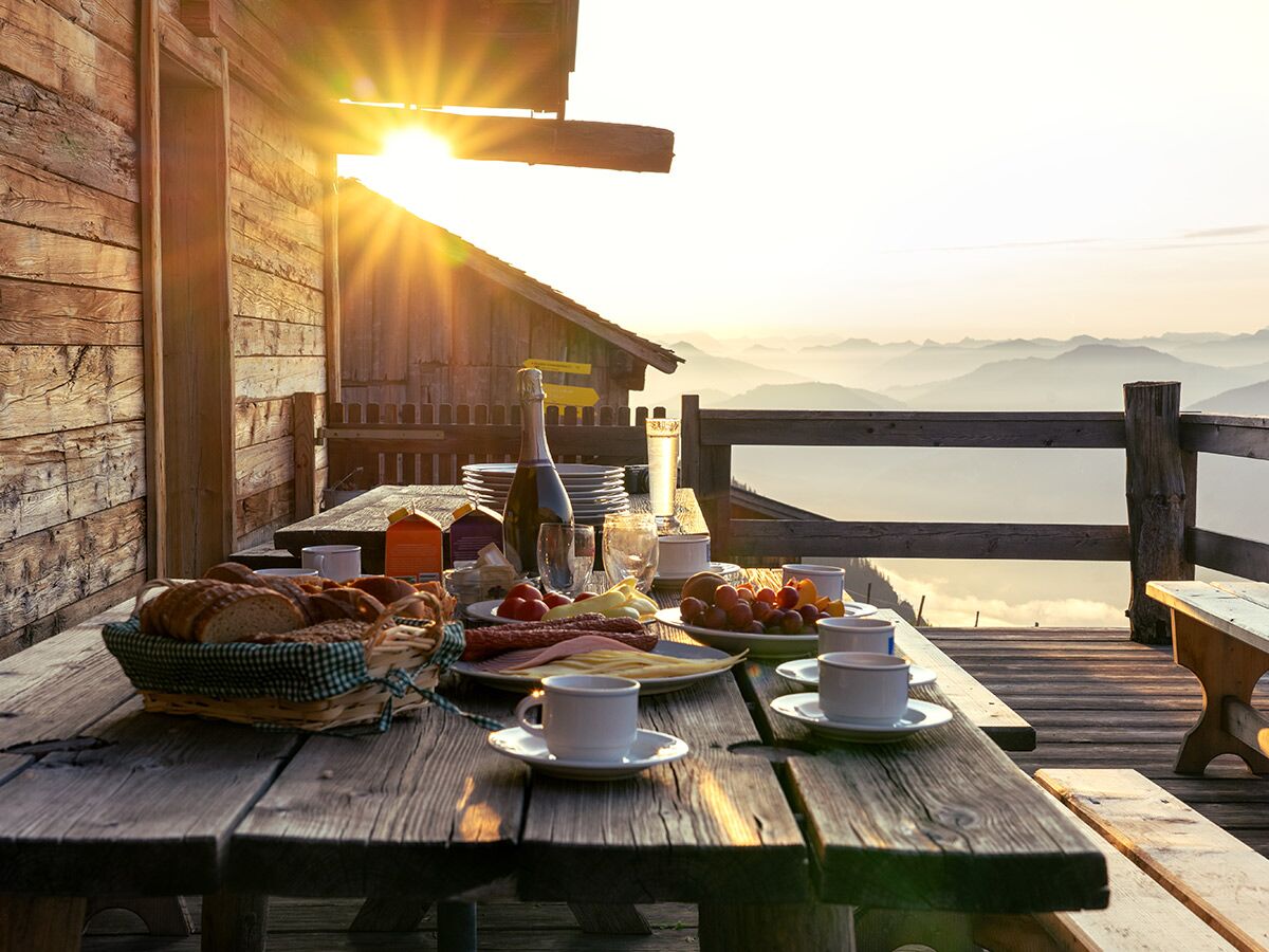 Gedeckter Frühstückstisch auf rustikaler Holzterrasse einer Berghütte bei Sonnenaufgang mit Blick auf die nebligen Alpen.