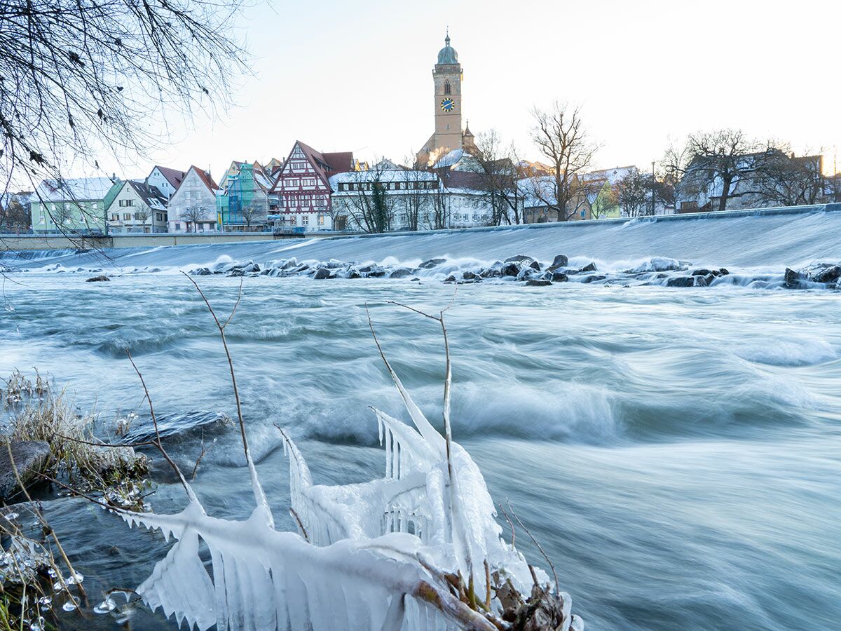 Winterlandschaft mit reißendem Fluss, Eiszapfen an Ästen im Vordergrund und einer malerischen Stadt mit Kirchturm im Hintergrund.