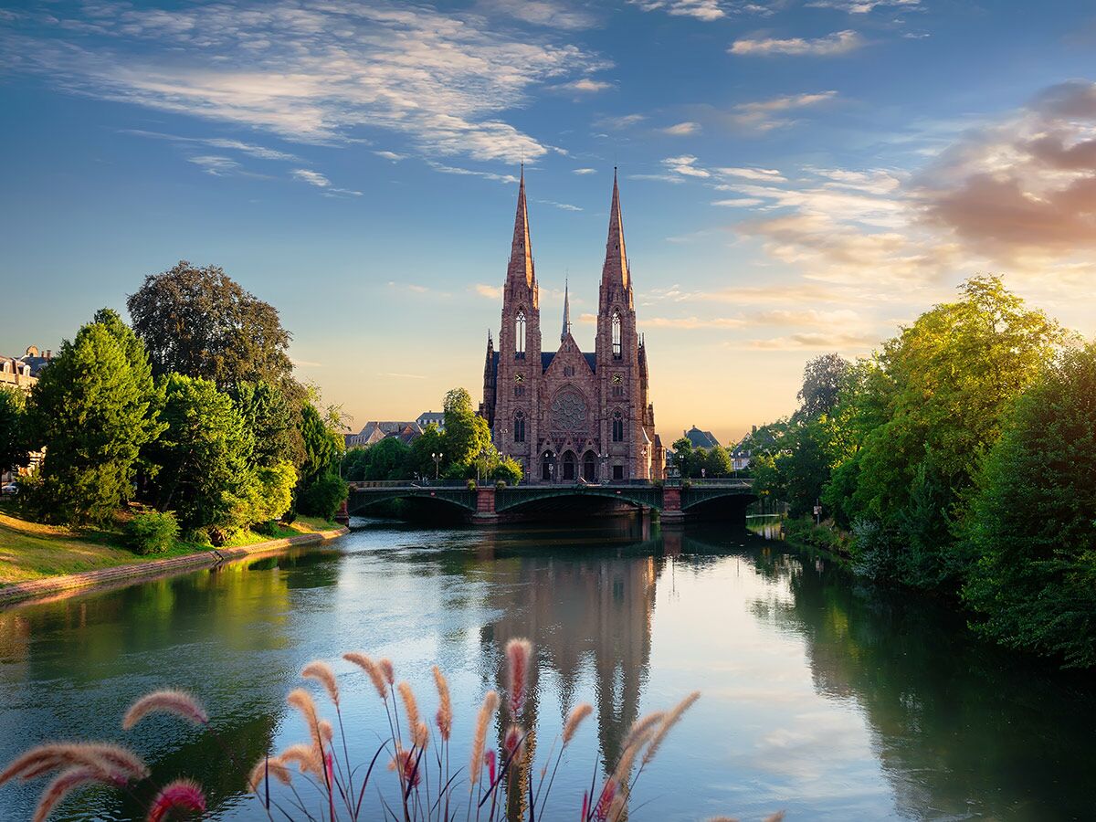 Die imposante Saint-Paul Kirche in Straßburg am Fluss Ill bei Sonnenuntergang mit Brücke und Spiegelungen im Wasser.