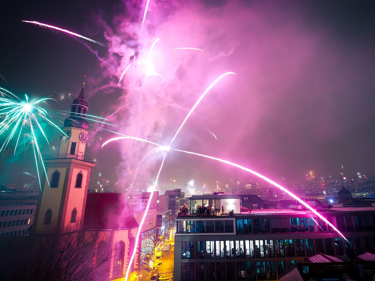 Nächtliche Stadtszene mit Glockenturm und pinkem, grünem Feuerwerk über Dächern, beleuchtet den Himmel festlich zur Feier.