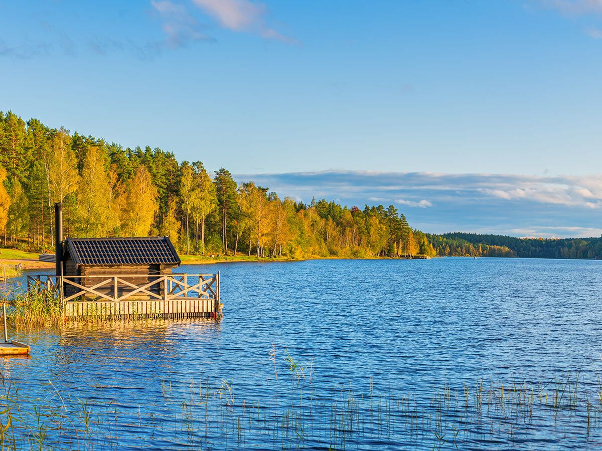 Holzsauna auf einem Steg am Seeufer, umgeben von einem bunten Herbstwald unter blauem Himmel. Ruhige skandinavische Landschaft.