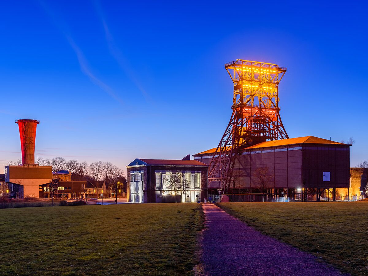 Beleuchtetes Fördergerüst einer Zeche bei Dämmerung mit rotem Wasserturm und modernen Gebäuden auf einem Industriegelände.