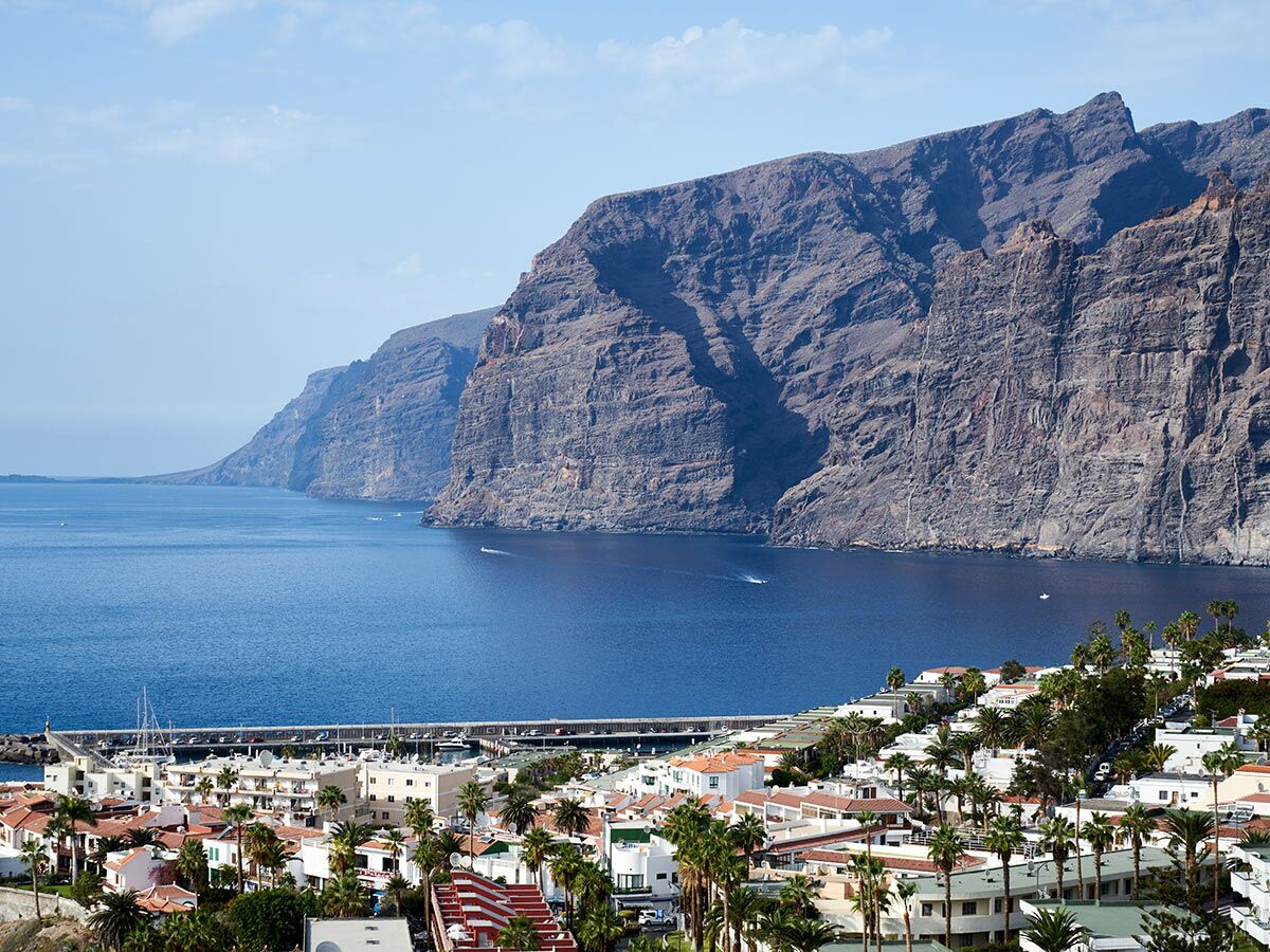 Panoramablick auf die Küstenstadt Los Gigantes auf Teneriffa mit Yachthafen, Häusern und imposanten Klippen am blauen Meer unter klarem Himmel.