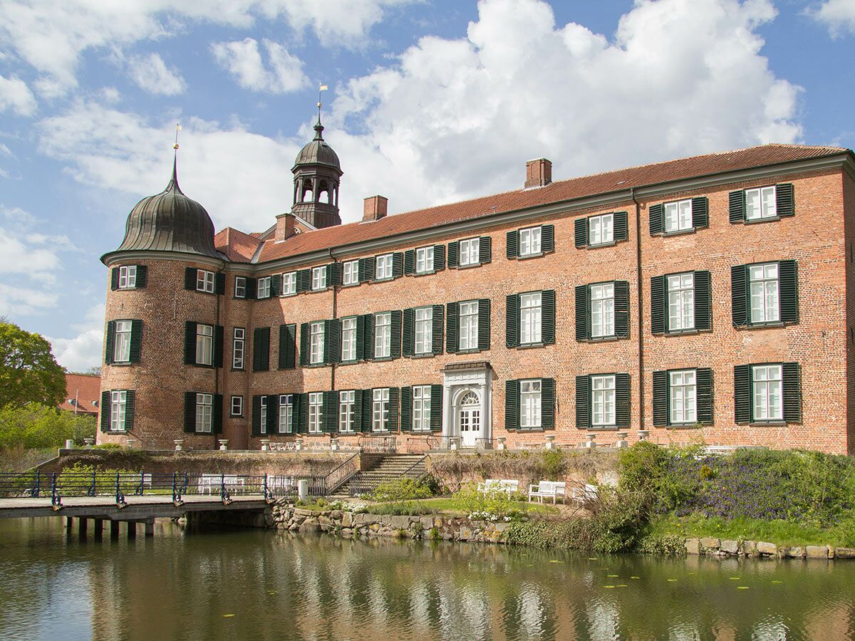 Historisches Backsteingebäude mit Türmen, grünen Fensterläden und einer Brücke über einen Wassergraben an einem sonnigen Tag.