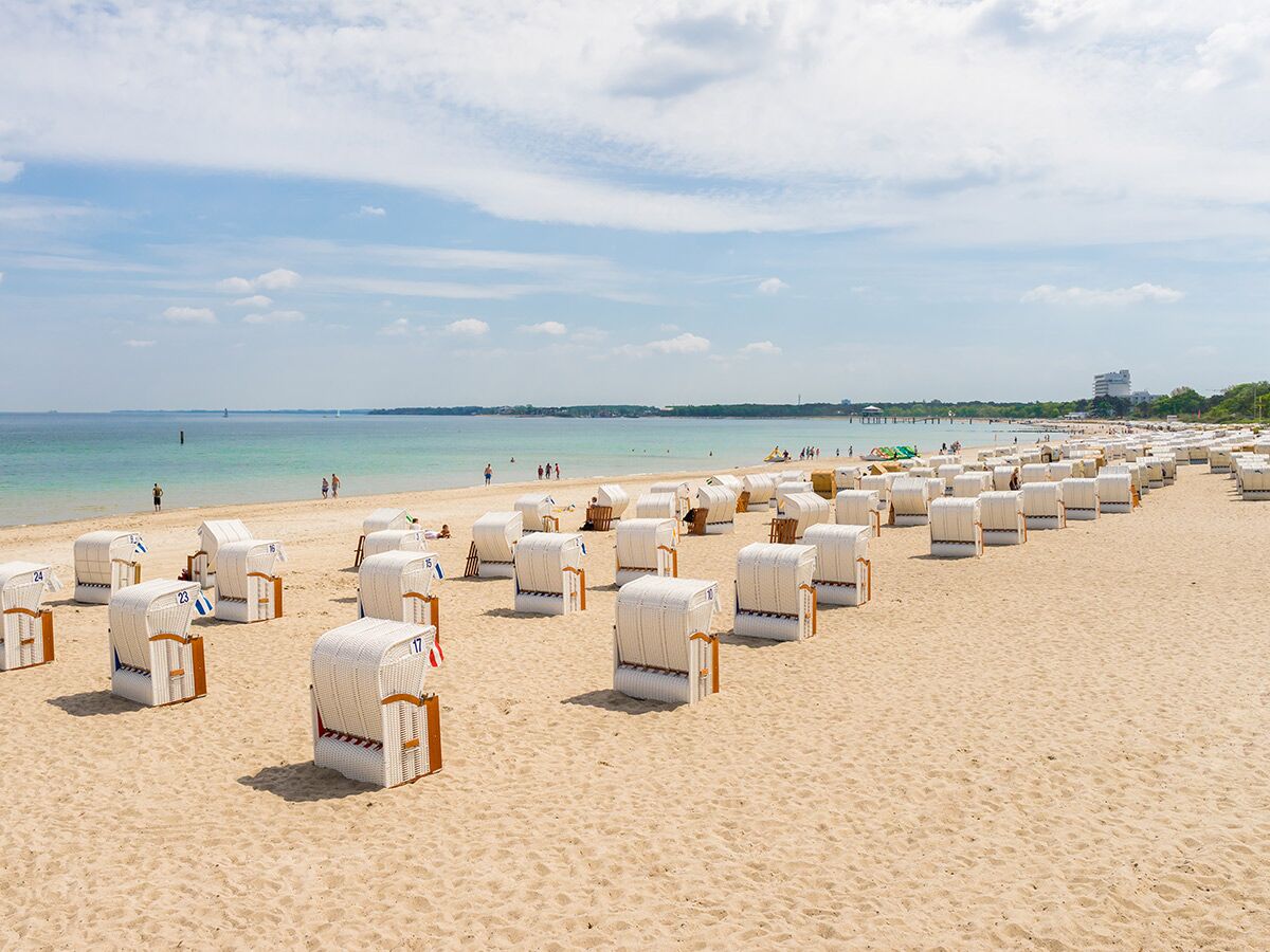 Sonnenbeschienener Sandstrand mit vielen weißen Strandkörben, Meer und Badegästen unter blauem Himmel.