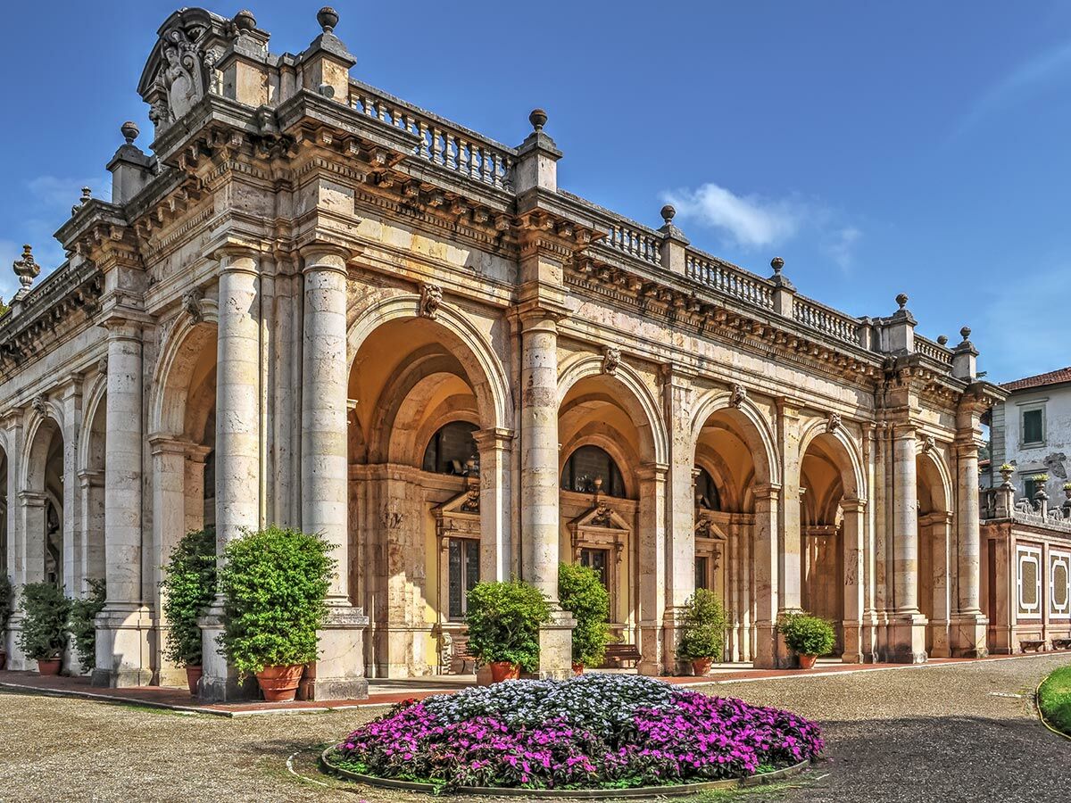 Ansicht einer eleganten historischen Loggia mit steinernen Säulenarkaden und einem farbenfrohen Blumenbeet im Vordergrund.