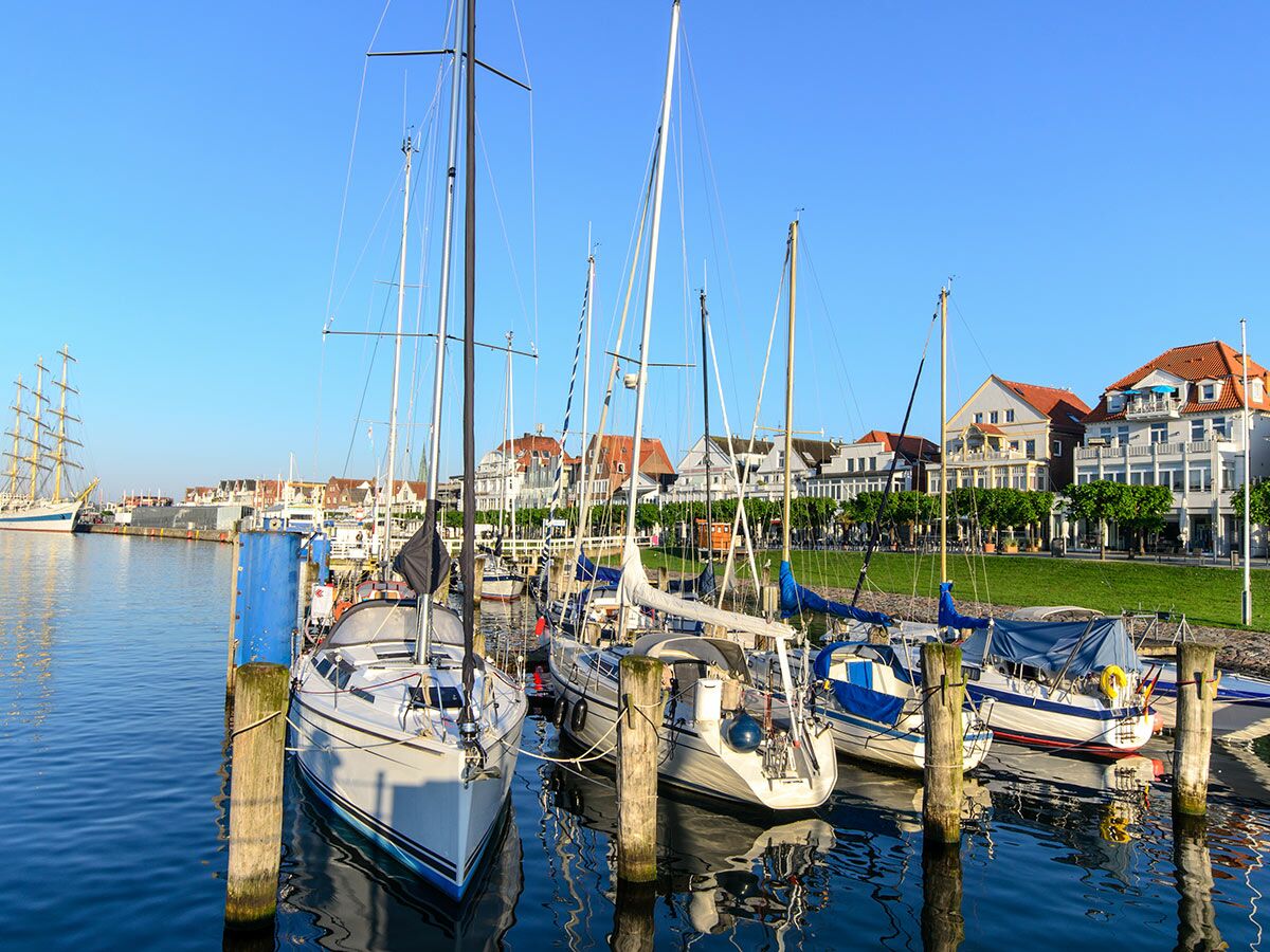 Segelboote liegen im sonnigen Hafen einer deutschen Küstenstadt mit traditionellen Gebäuden und einem Großsegler im Hintergrund.