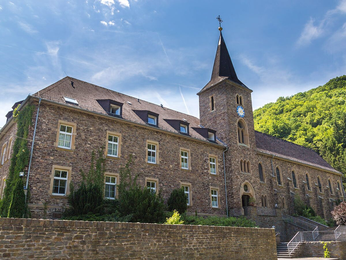 Historisches Kloster oder Abtei aus Stein mit hohem Kirchturm und Uhr, umgeben von grünen Hügeln unter blauem Himmel.
