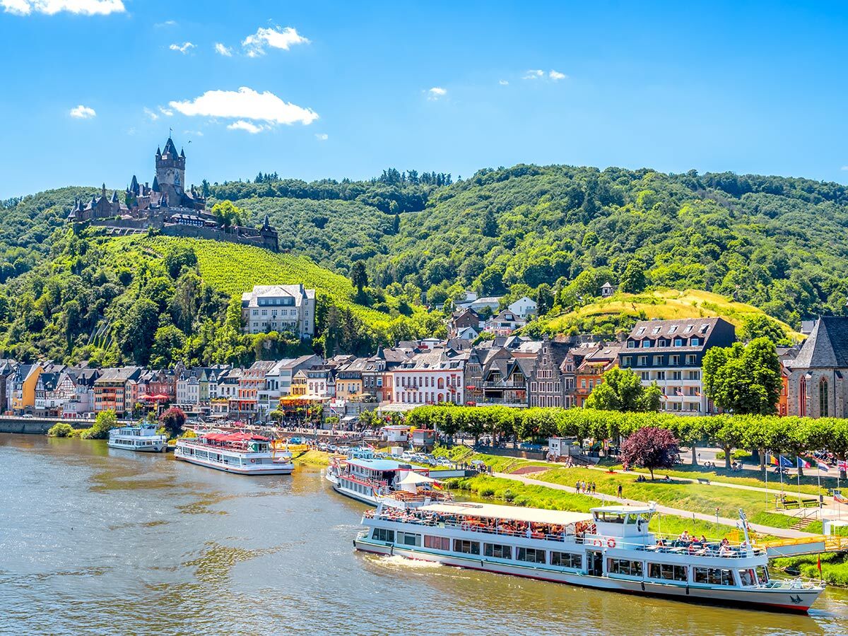 Panoramablick auf Cochem mit Reichsburg, Mosel, Schiffen und bunten Häusern am sonnigen Flussufer.