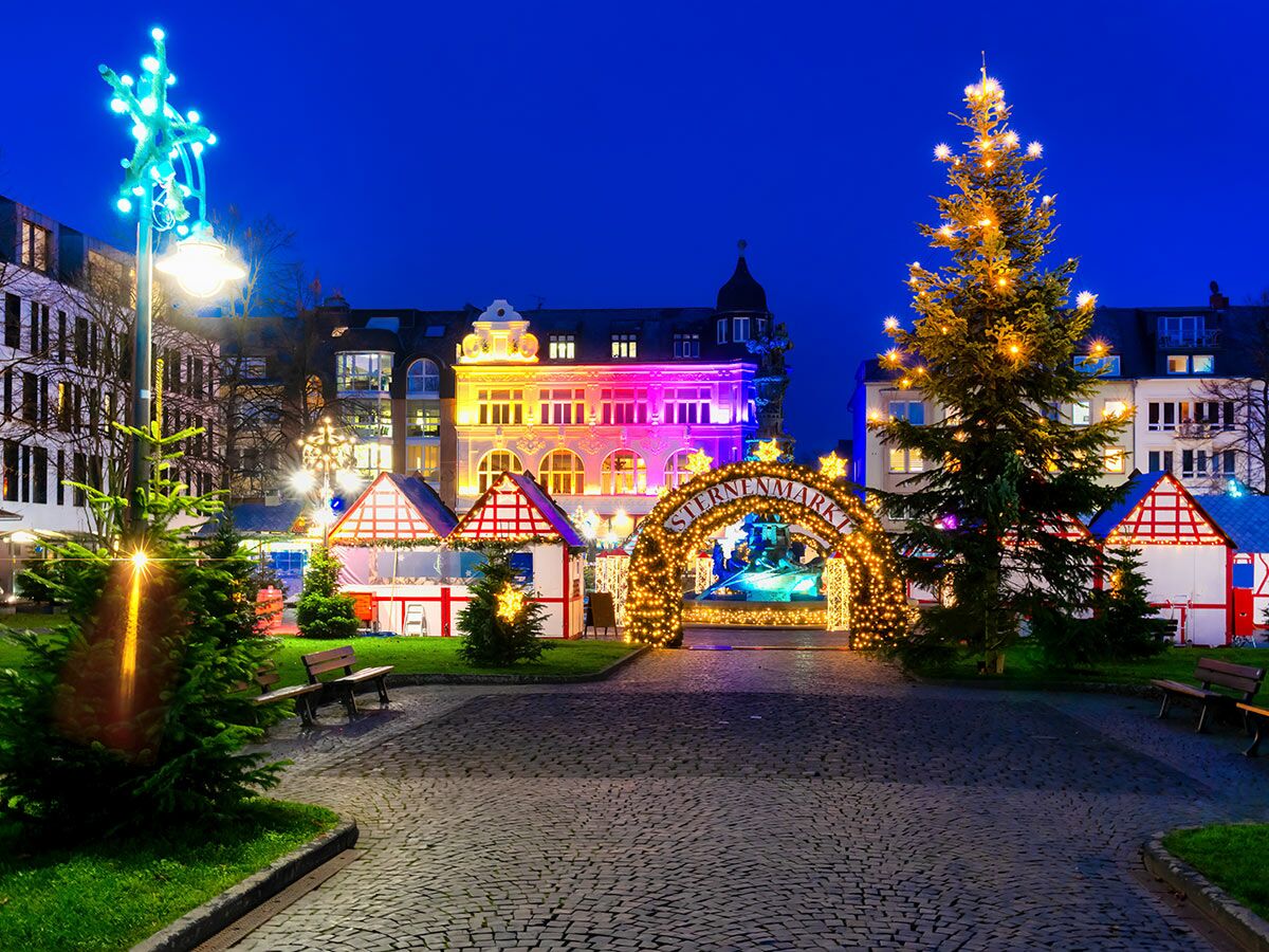 Beleuchteter Sternenmarkt am Abend mit festlich geschmückten Weihnachtsbäumen, traditionellen Holzbuden und einem Lichterbogen.