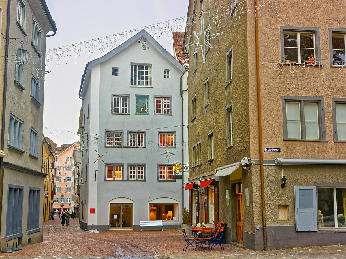 St. Martinsplatz, Chur, Schweiz: Historische Gasse mit Gebäuden, Weihnachtsbeleuchtung, Café und Rembrandt-Gemälde im Winter.