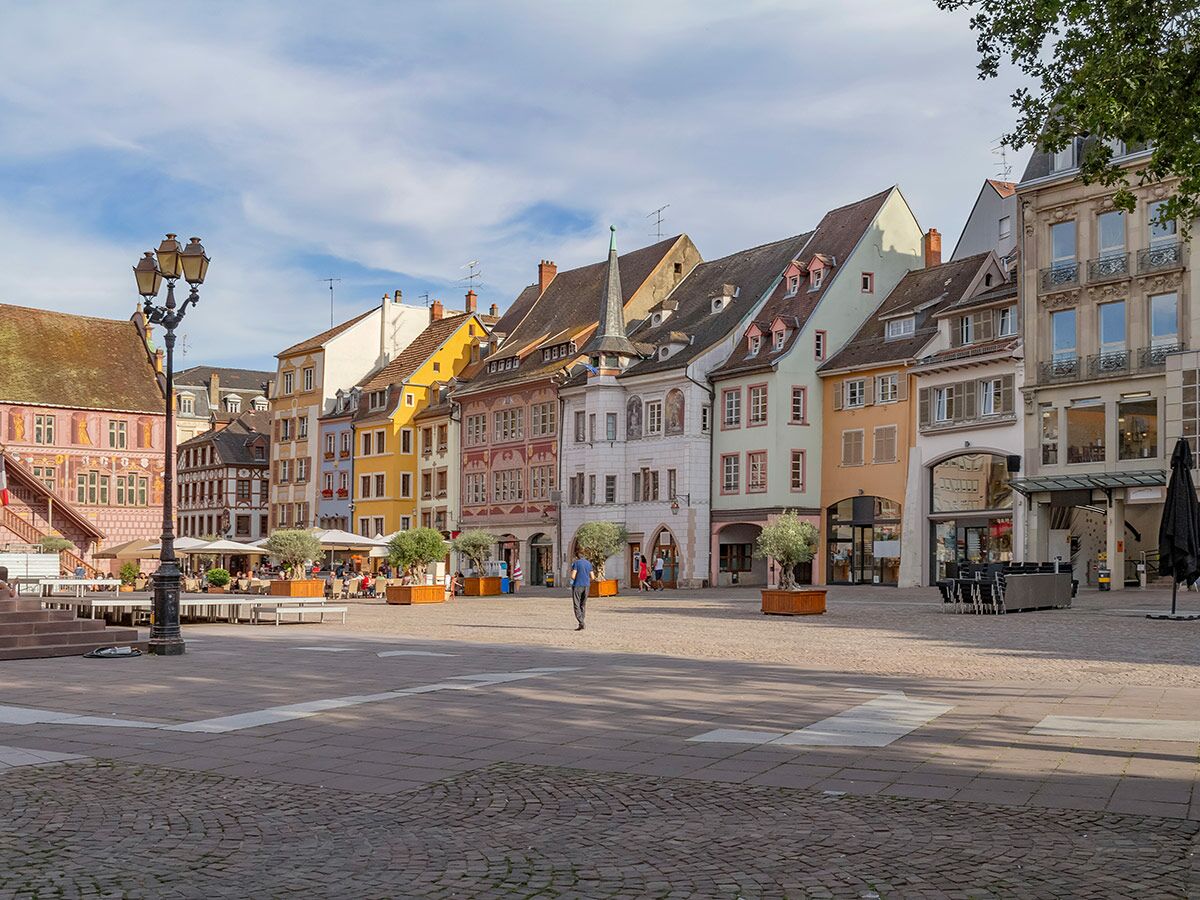 Sonniger europäischer Marktplatz mit einer Reihe bunter, historischer Gebäude, Straßencafés und Menschen.