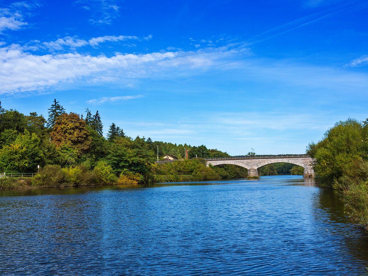 Malerische Flusslandschaft mit alter Steinbrücke, umgeben von grünen und herbstlich gefärbten Bäumen unter blauem Himmel.