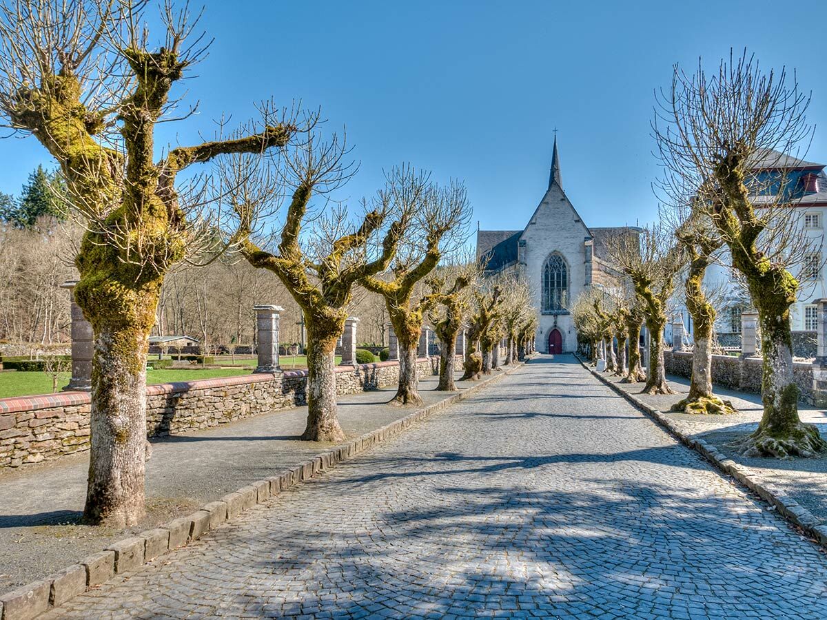 Lange gepflasterte Allee führt zu einer historischen Abteikirche mit spitzem Turm unter blauem Himmel, flankiert von moosbewachsenen Bäumen.