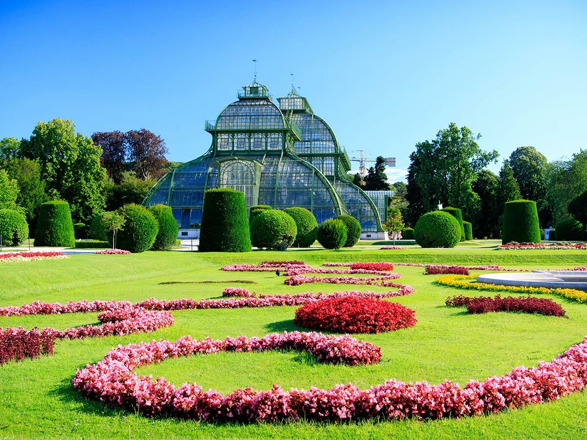 Panoramablick auf das historische Palmenhaus in Schönbrunn, umgeben von gepflegten Rasenflächen und farbigen Blumenbeeten.