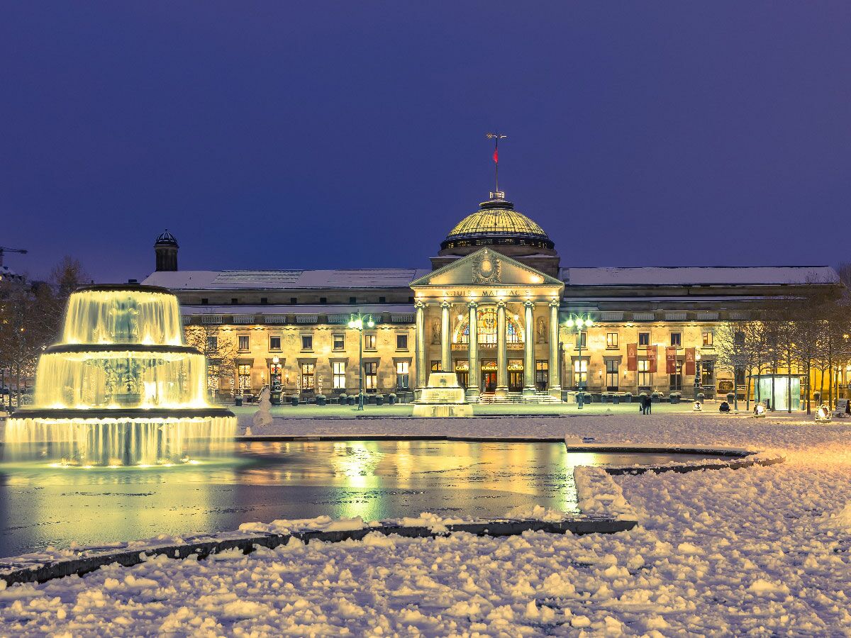 Beleuchtetes Kurhaus Baden-Baden bei Nacht im Winter, mit Schnee und leuchtendem Brunnen. Elegante historische Architektur.