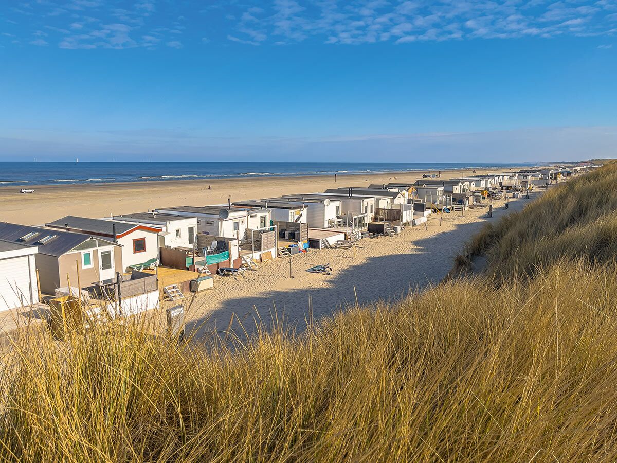 Blick über eine lange Reihe von weißen Strandhäusern am Sandstrand, dahinter das Meer und Dünen unter blauem Himmel.