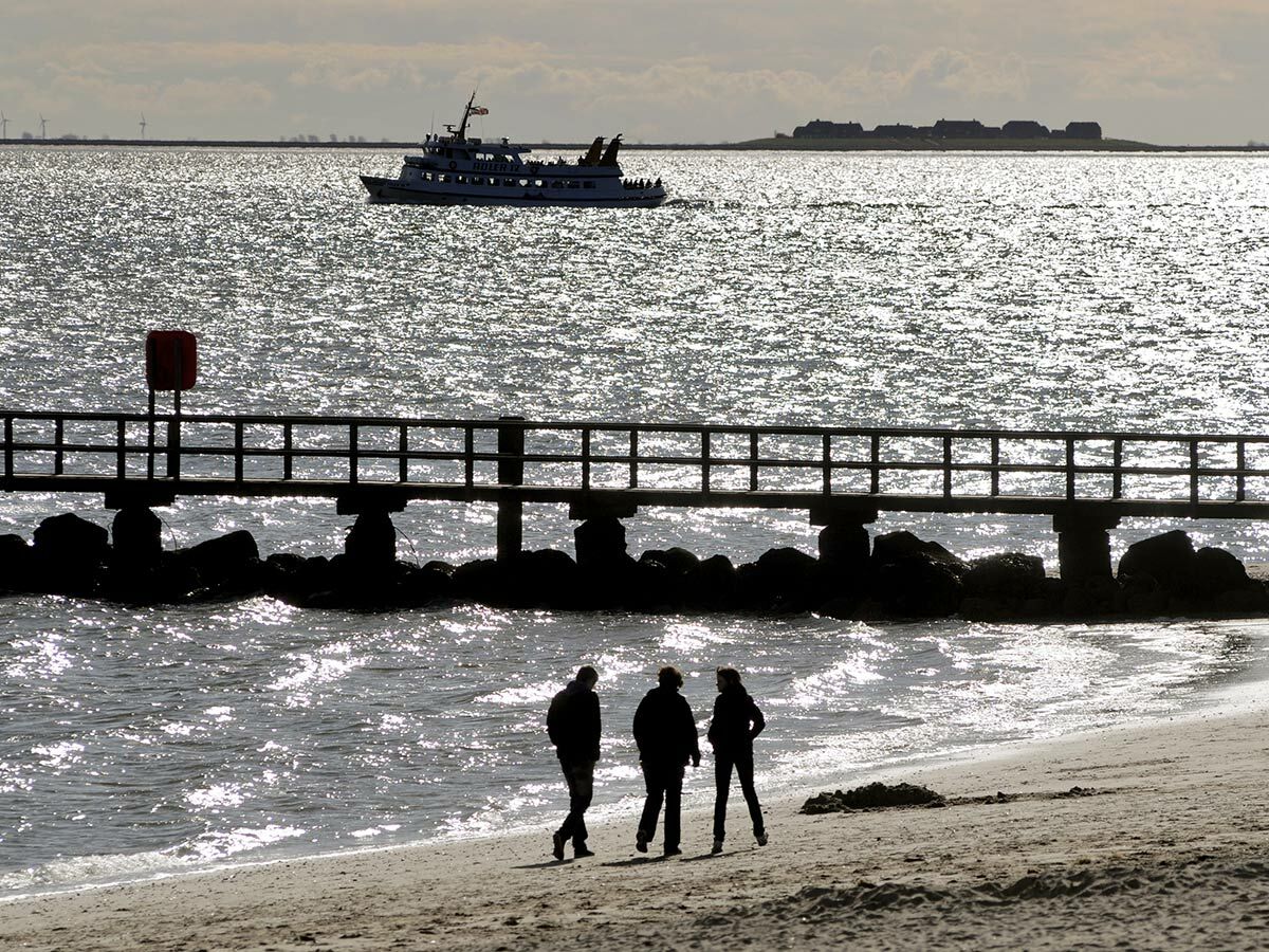 Drei Personen spazieren am Strand im Gegenlicht, mit Pier, glitzerndem Wasser, Boot und Insel im Hintergrund.