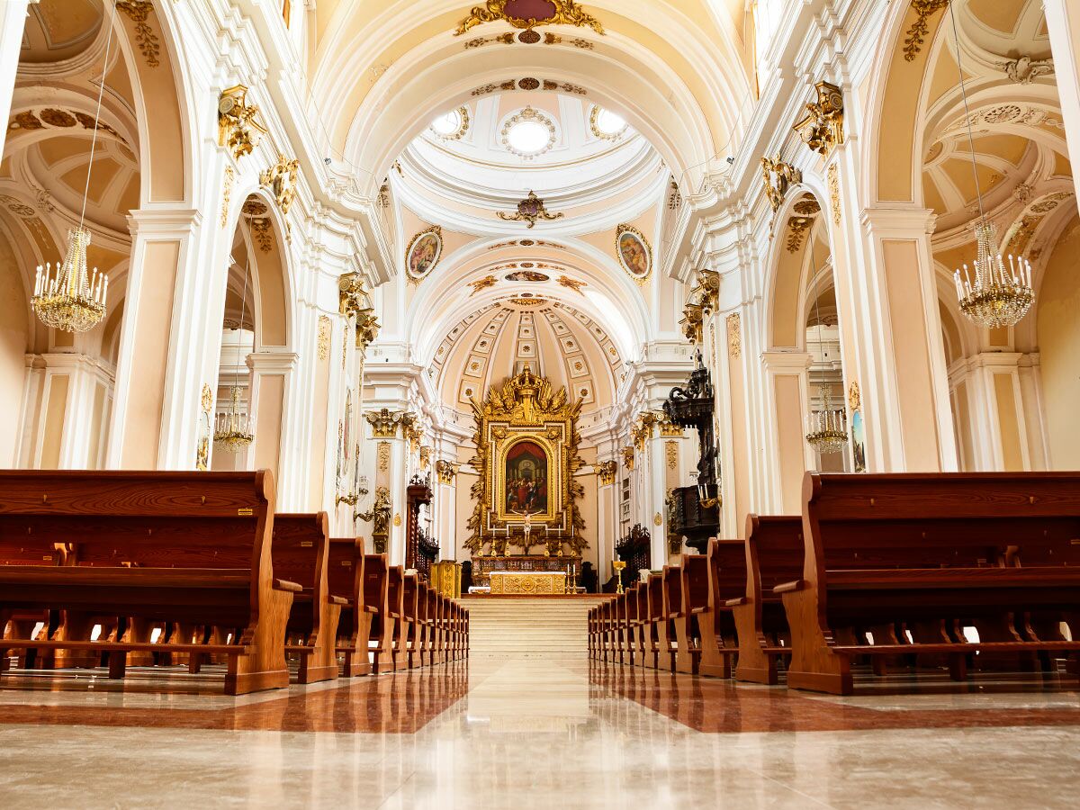 Innenansicht eines opulenten Kirchenschiffs mit Reihen von Holzbänken, hohen Gewölben und einem prächtigen Altar am Ende.