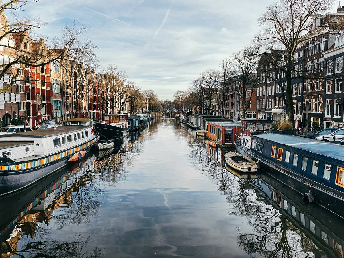 Blick auf einen Amsterdamer Kanal mit bunten Hausbooten, historischen Grachtenhäusern und Baumspiegelungen im Wasser unter bewölktem Himmel.