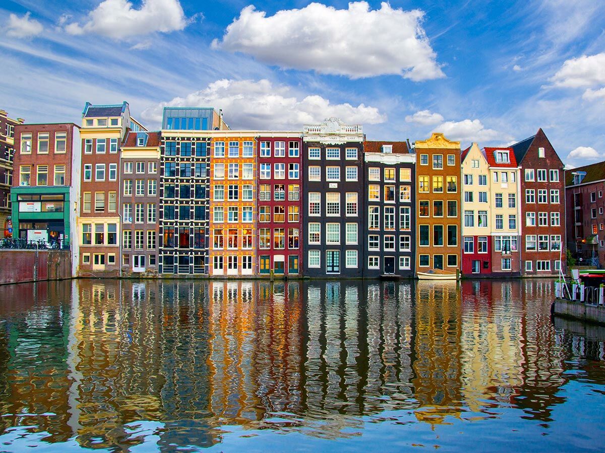 Reihe bunter Grachtenhäuser am Kanal in Amsterdam unter blauem Himmel, ihre Spiegelung im Wasser.