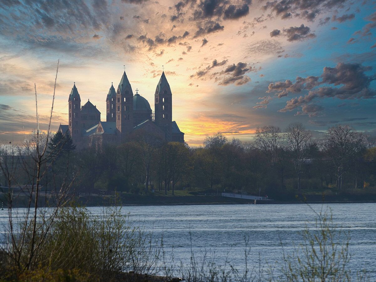 Majestätischer Speyerer Dom bei Sonnenuntergang am Rhein, umgeben von Bäumen und dramatischer Himmelsfärbung.