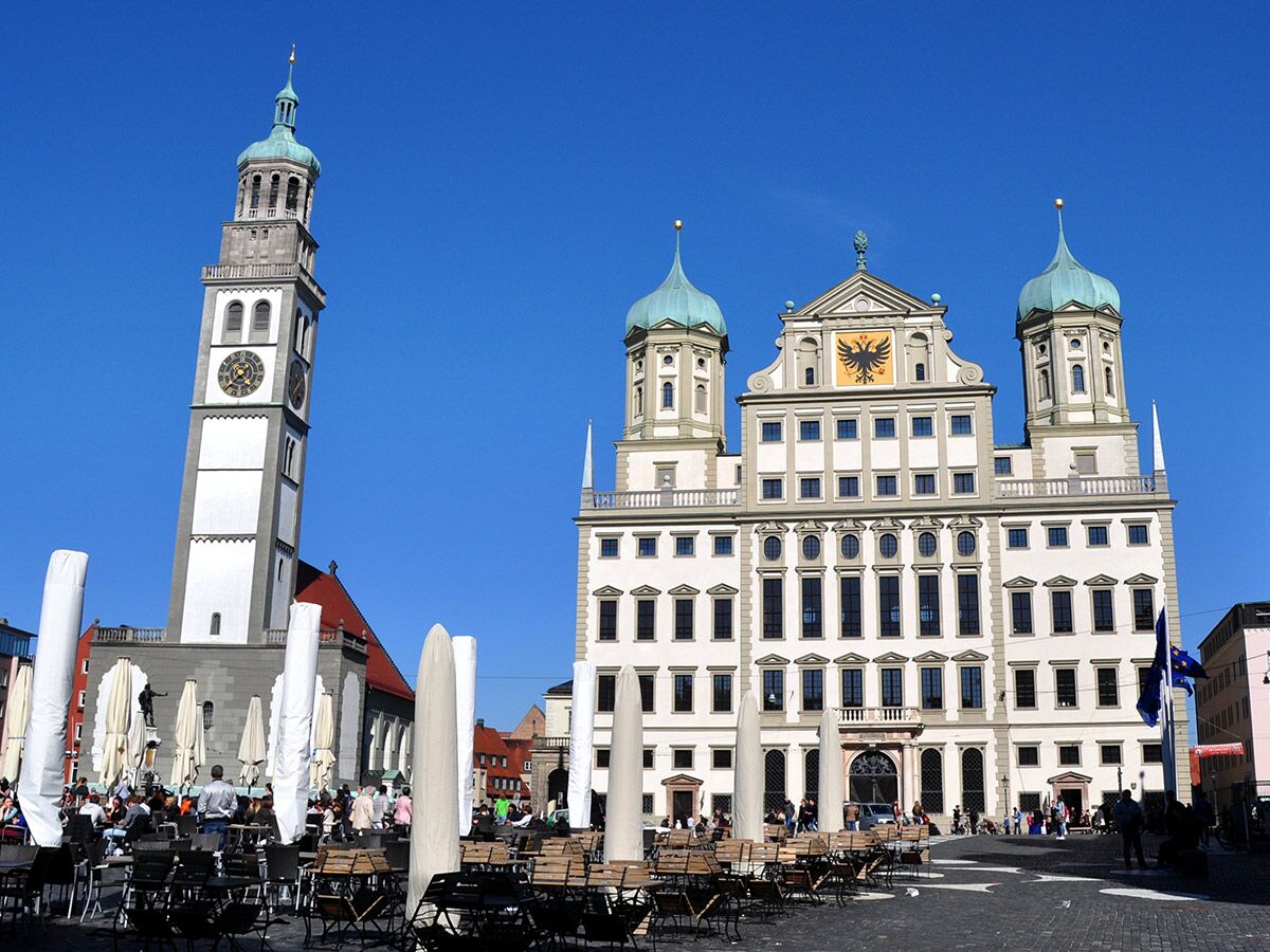 Blick auf den Augsburger Rathausplatz mit dem Perlachturm und dem historischen Rathaus unter blauem Himmel, Menschen auf Café-Terrassen.